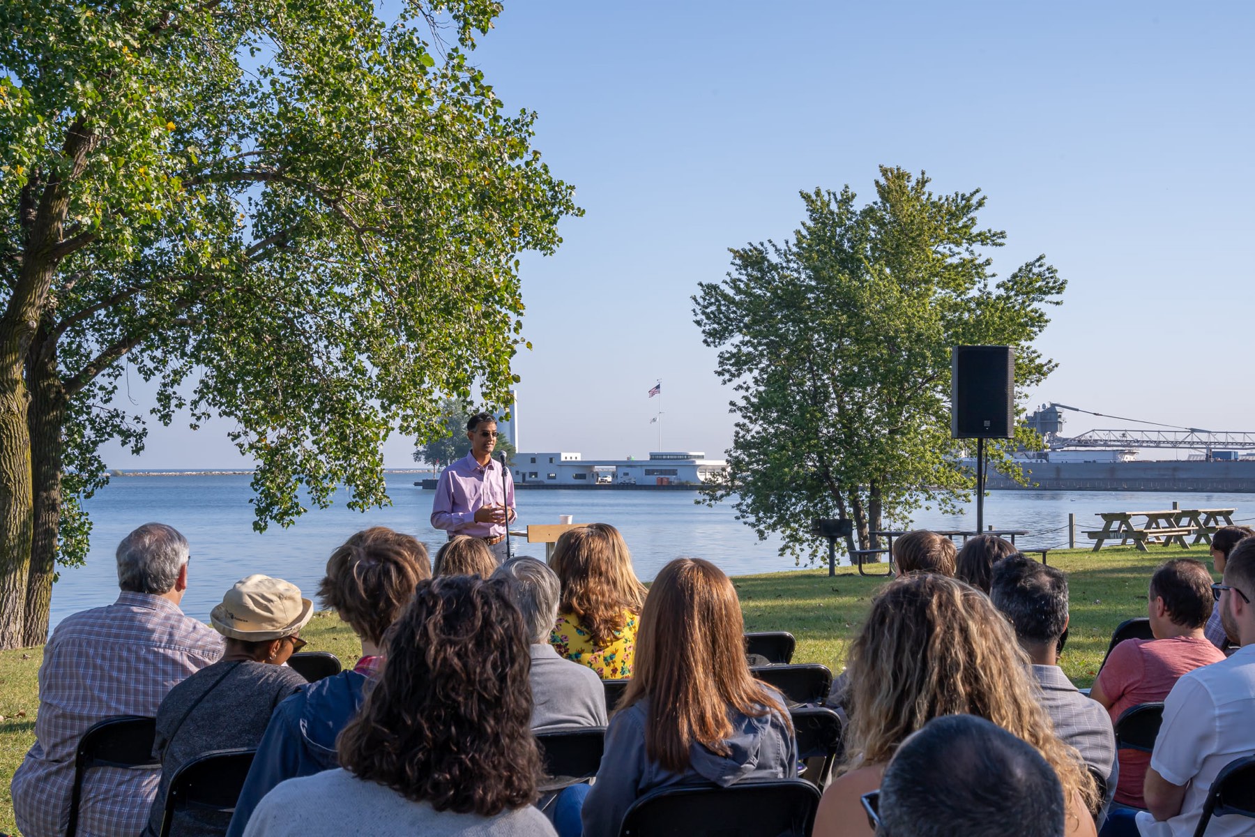 An audience on folding chairs listening to a talk on the lawn at Wendy Park, Lake Erie behind them