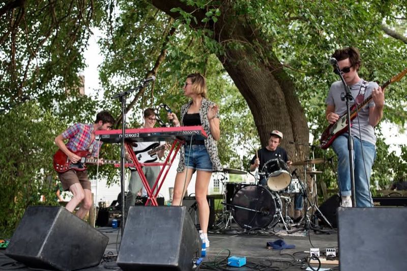 A band with keyboard, guitars and drums plays under a big tree.